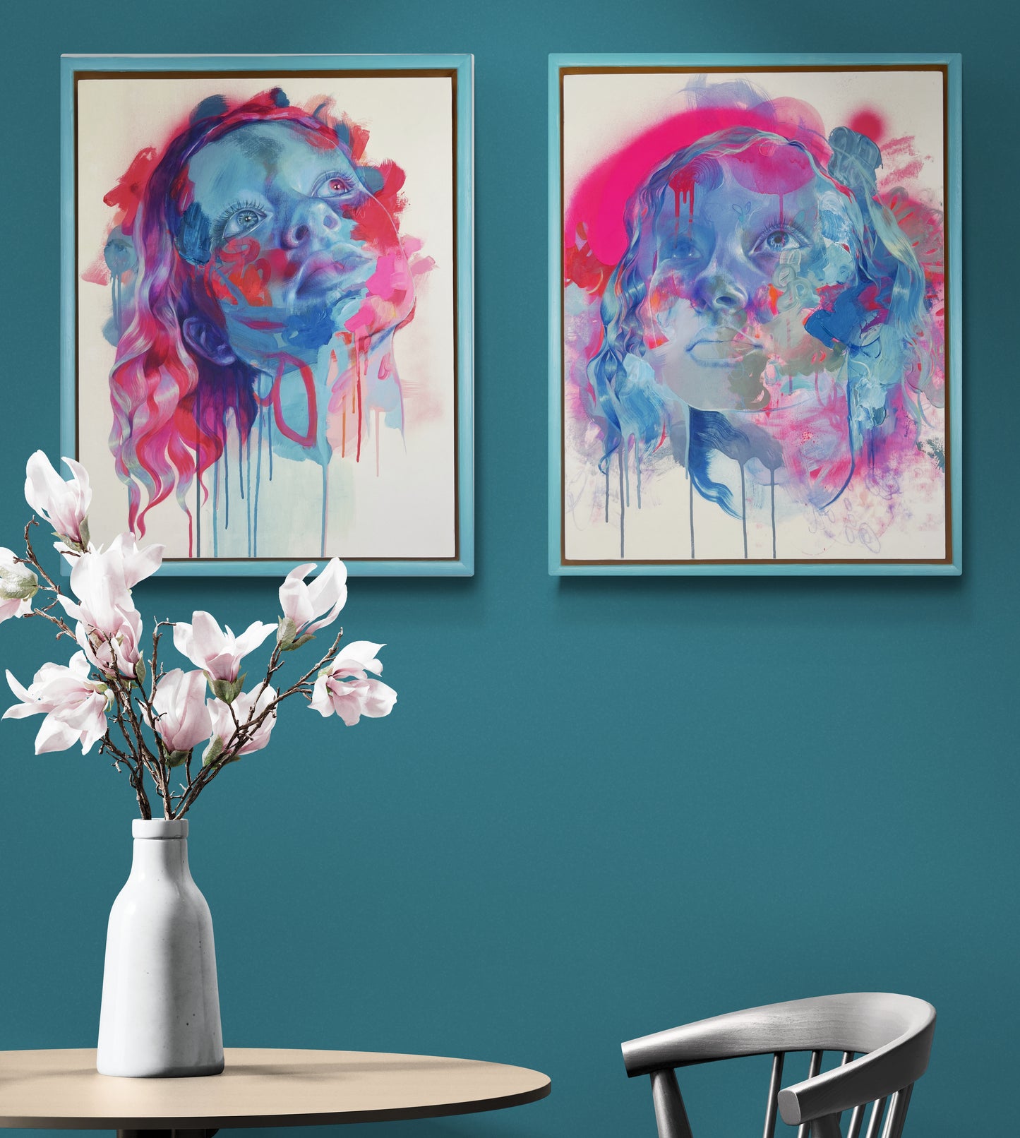 Picture of pair of framed artworks hanging on green background, above table w=and chair with vase of flowers on left